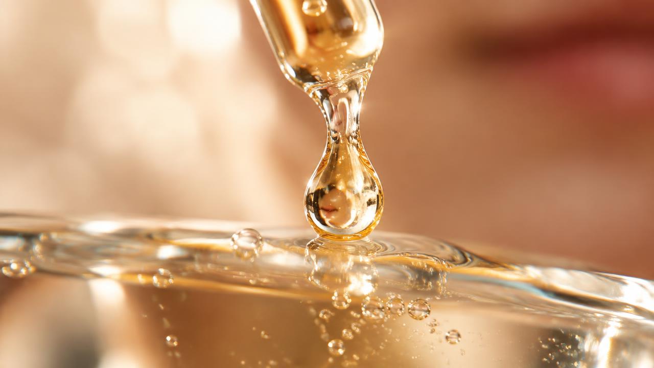 Backlit macro photograph of a golden cosmetic serum drop touching down on a glass surface